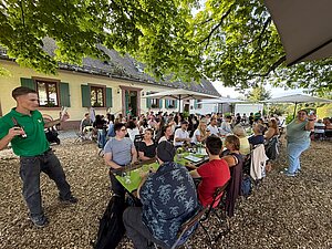 Kolleg:innen sitzen und stehen bei geselligem Beisammensein vor dem Gebäude des Hofguts im Schatten großer Bäume.